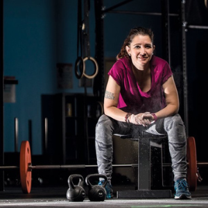 Nicki Perkins sitting on a bench smiling in a CrossFit gym with weights behind her.